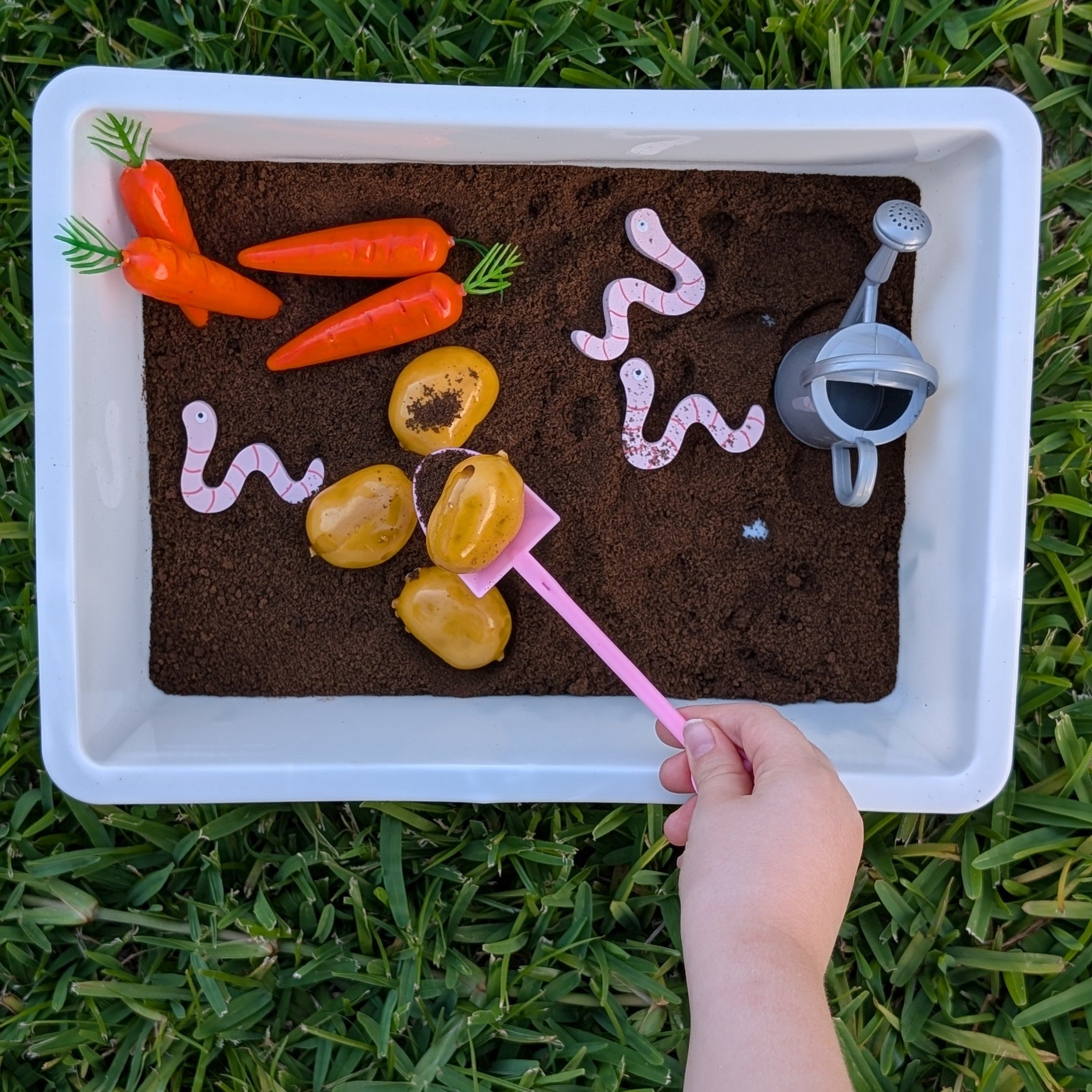Sensory bin with soil, vegetables, and a pink tool on grass
