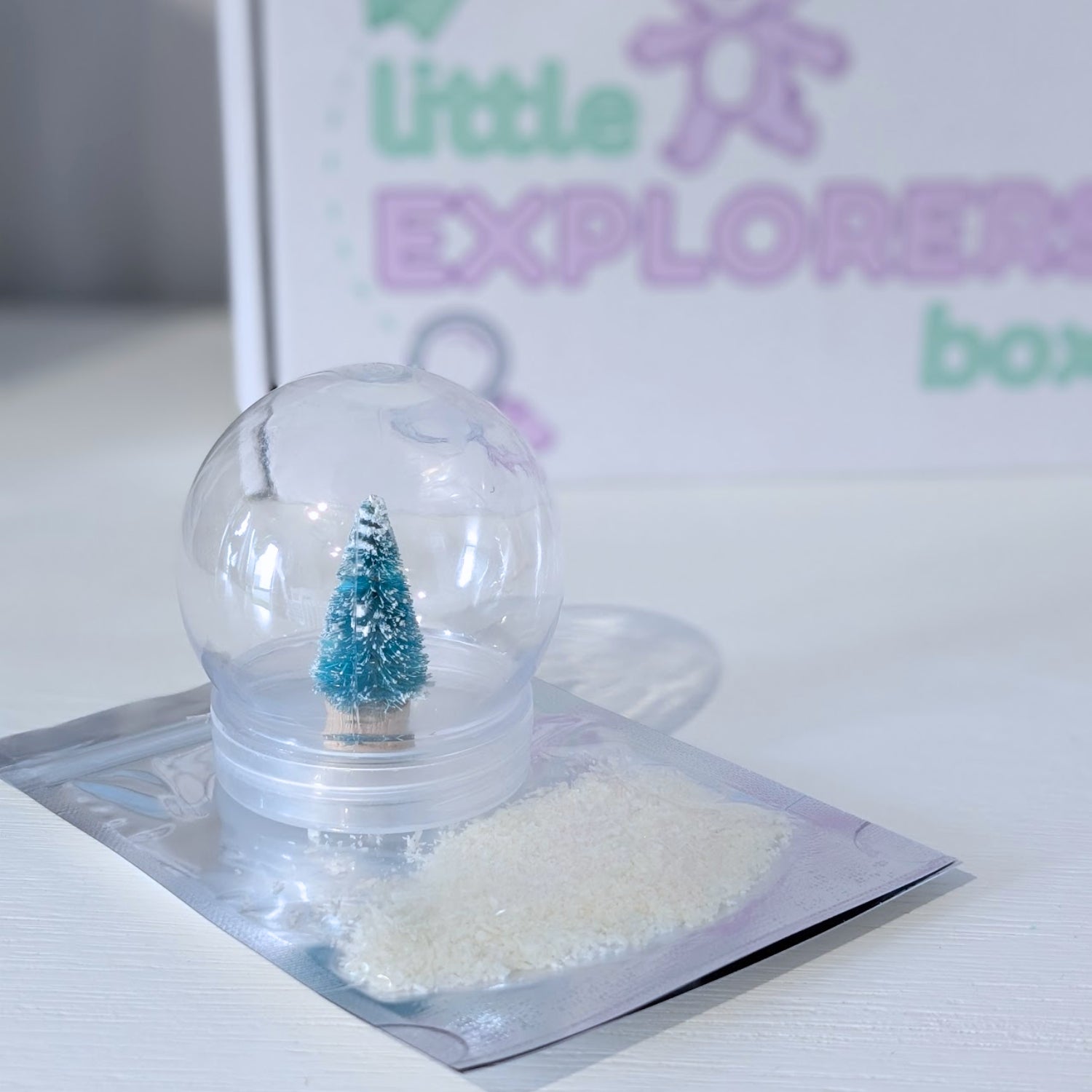 Small snow globe with a miniature tree inside, placed on a white surface with a 'Little Explorers' box in the background.
