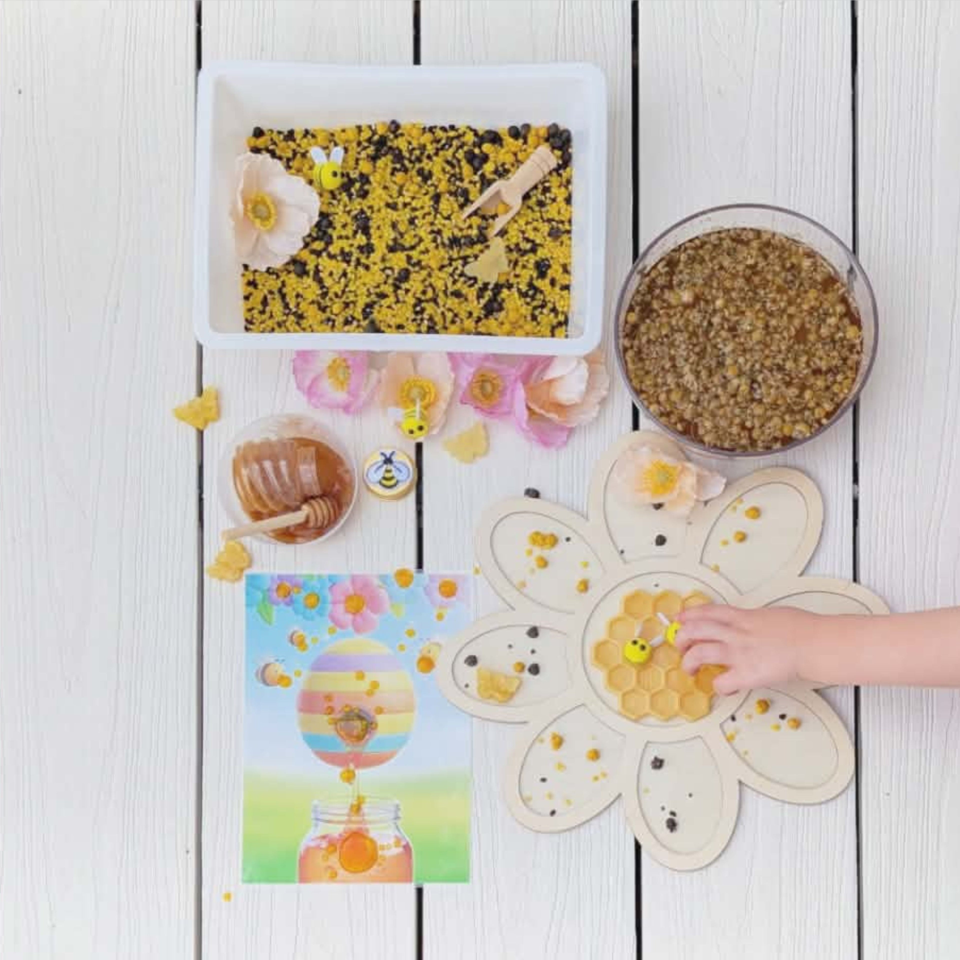 Children's sensory play activity box with four engaging activity laid out on the floor. The activities include a flower petal potion, a dry sensory mix with black and yellow textures, felt bees and resin butterflies, a pot of honey and spoon for a messy play adventure and a laminated picture of a bee hive for another play activity. 