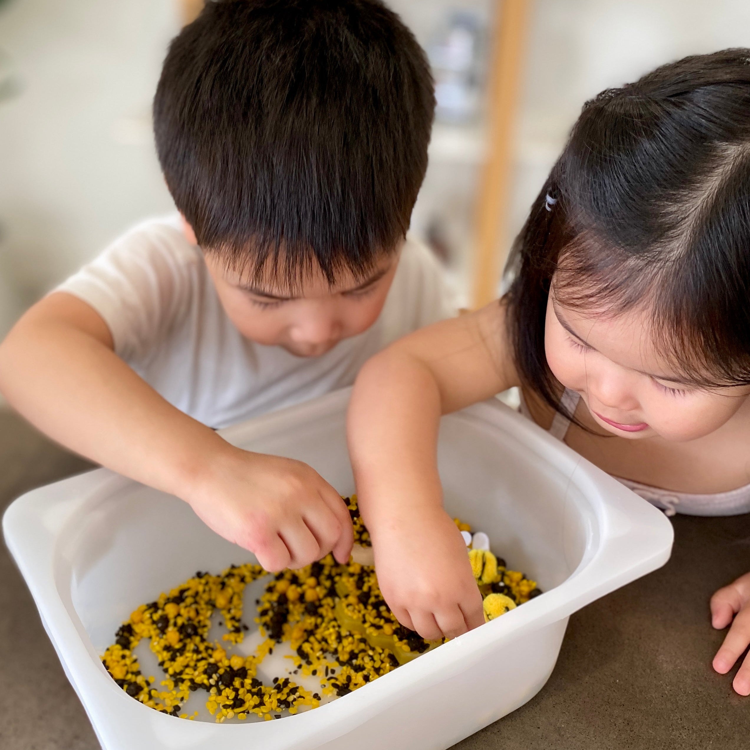 Two children sitting at the table playing with yellow and black dry sensory treasures in a white container with little felt bee toys. 