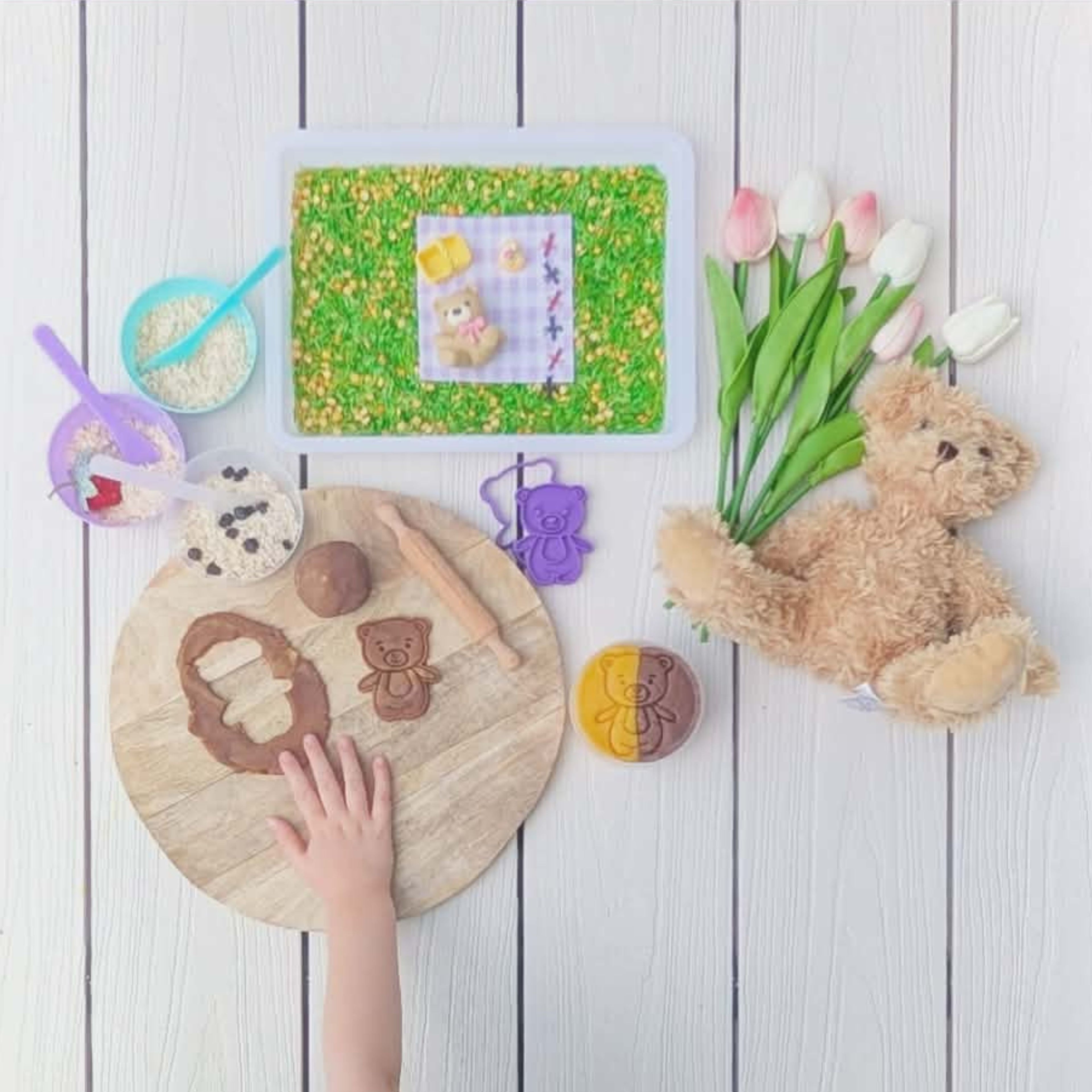 Children's sensory play activity box with three engaging activity laid out on the floor. The activities include a dry tray play tray with a sensory base that represents grass and flowers, a teddy bear and picnic rug, picnic basket and snacks and some cheeky ants out to invade the picnic. The next activity is Brown Bear & Honey Dough to press and cut out some little bear friends. The final activity is based on goldilocks and the three bears for some book ish play making the three bears porridge. 
